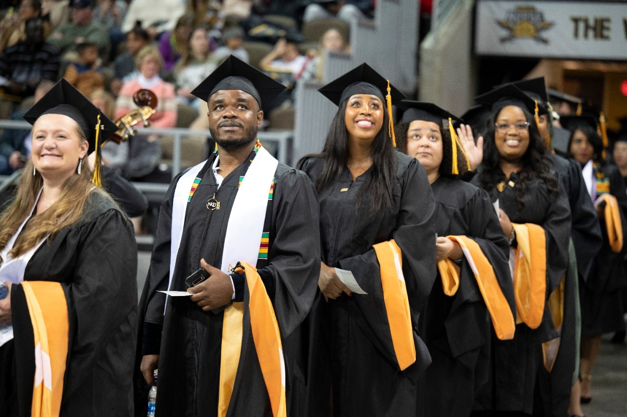 A line of students enter Truist Standium for the Fall 2022 commencement ceremony. Three black students look up at the families in the stands. 