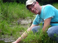 Photo of Dr. Cooper kneeling in the grass at the side of a pond, taking a sample of the water.