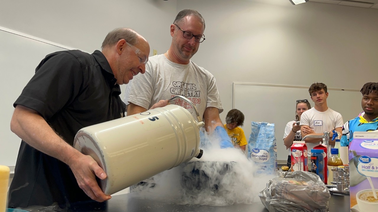 Beekers filled with colorful water sit on a table with elementary children in the background.
