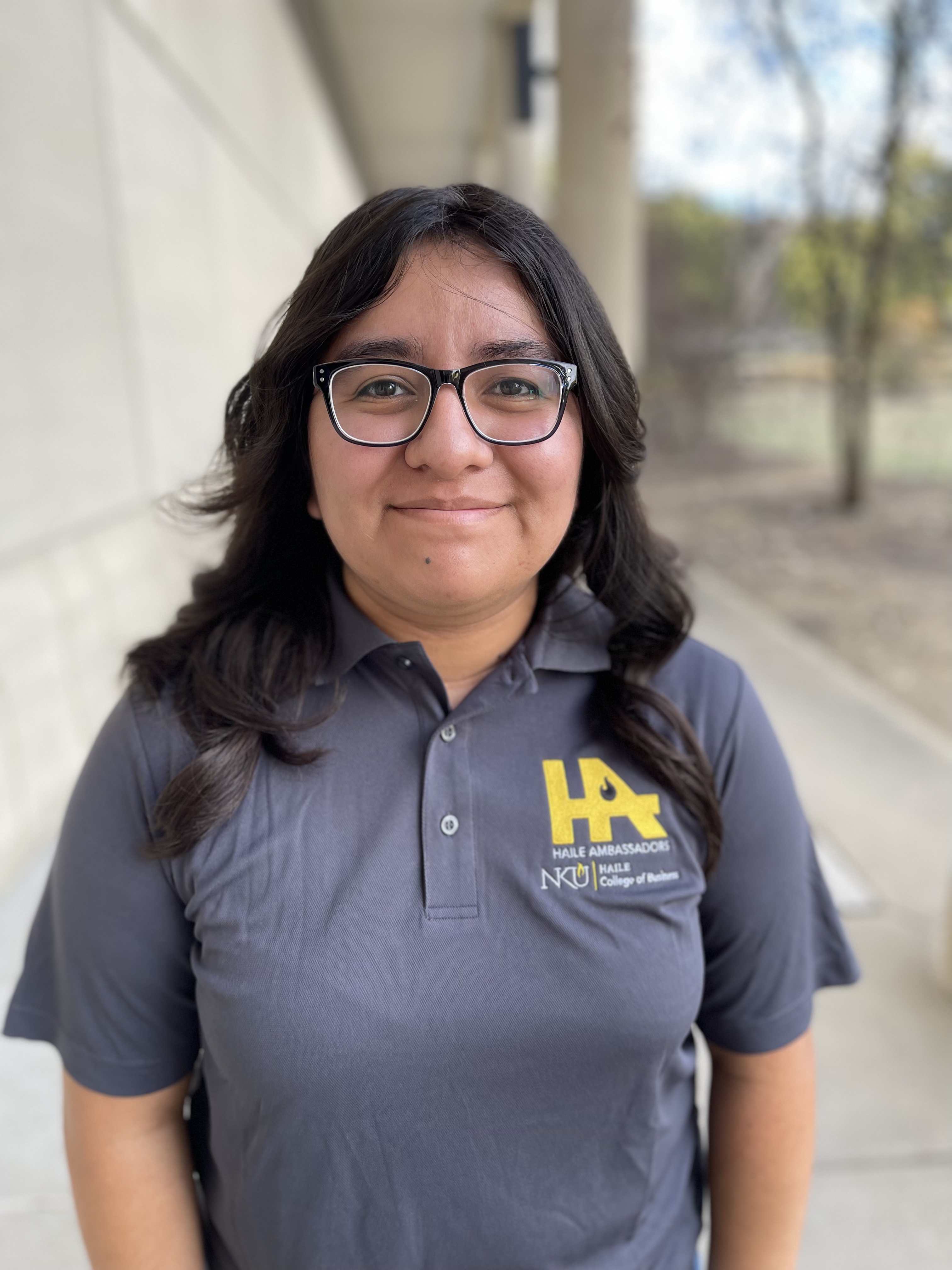 Janeth Vazquez-Hernandez smiles while standing outdoors near a columned building. She has long dark hair, is wearing glasses, and a gray NKU Haile Ambassadors polo shirt.