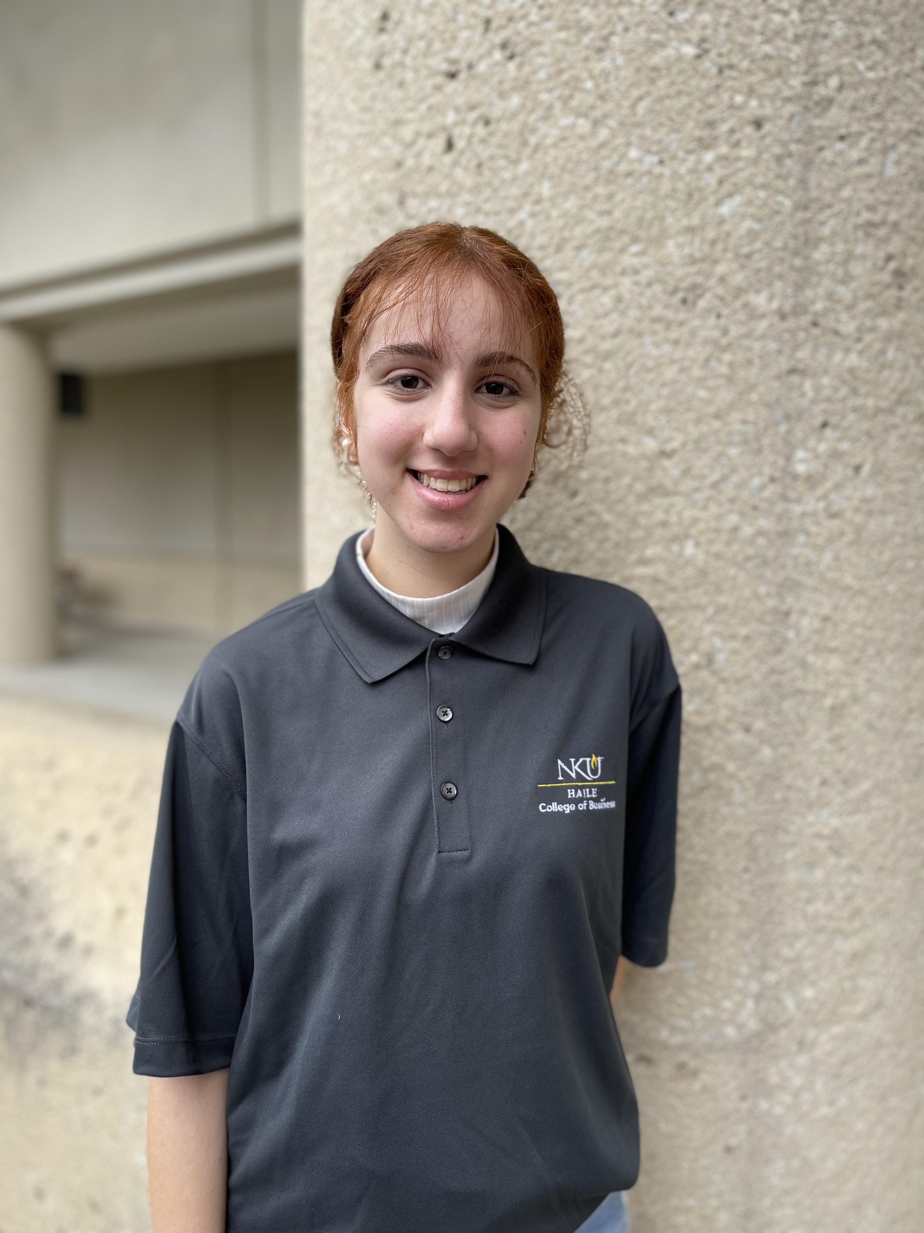 Sandra Saba smiles while standing outdoors in front of a textured column. She has red hair pulled back and is wearing a gray NKU Haile College of Business polo shirt.