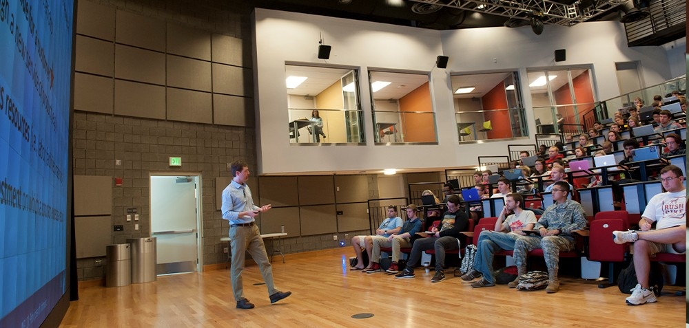 Professor speaking in front of the Digitorium screen showing the audience in seating.