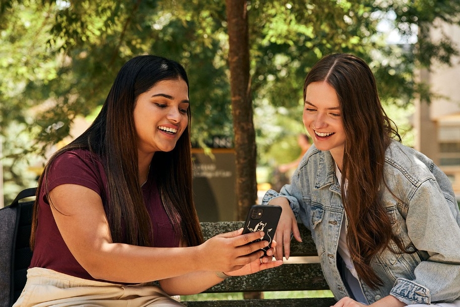 Students sitting outside and smiling while looking at pictures on a phone