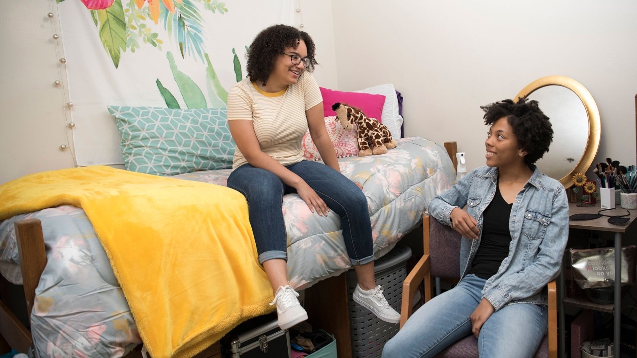 Two roommates sitting in their NKU dorm room, smiling at each other.