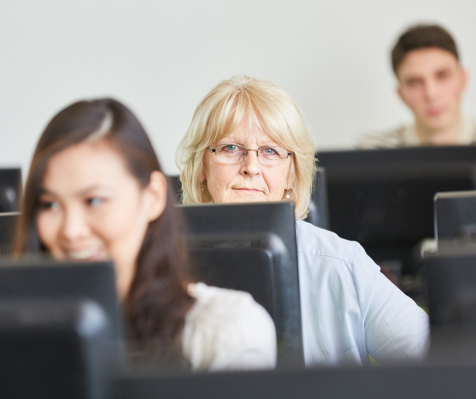 An older woman looks over a computer monitor in a classroom.