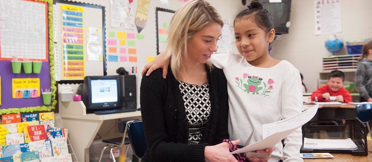 Teacher sits with a student in a brightly decorated elementary school classroom.