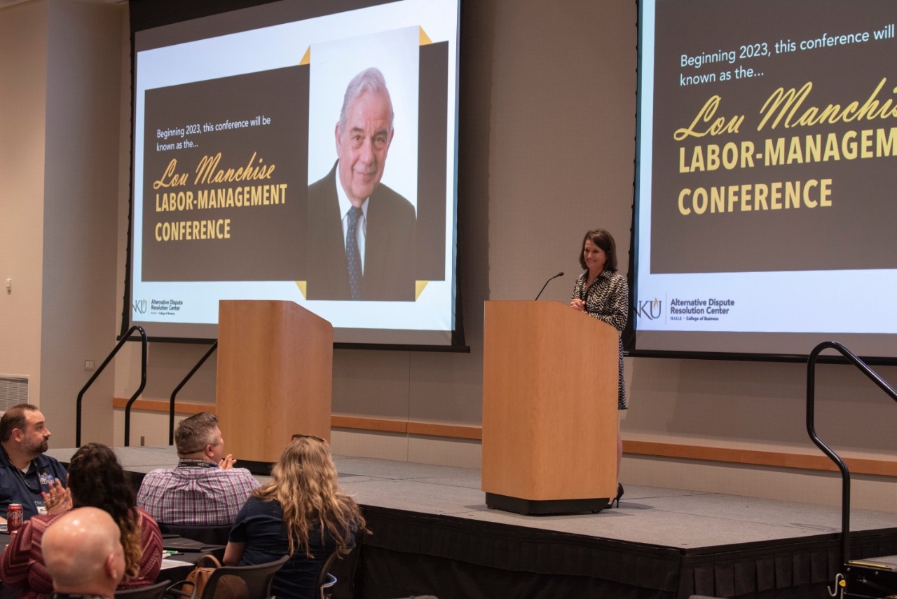 A woman stands behind a podium in front of a powerpoint presentation about the Loud Manchise Labour Management Conference.
